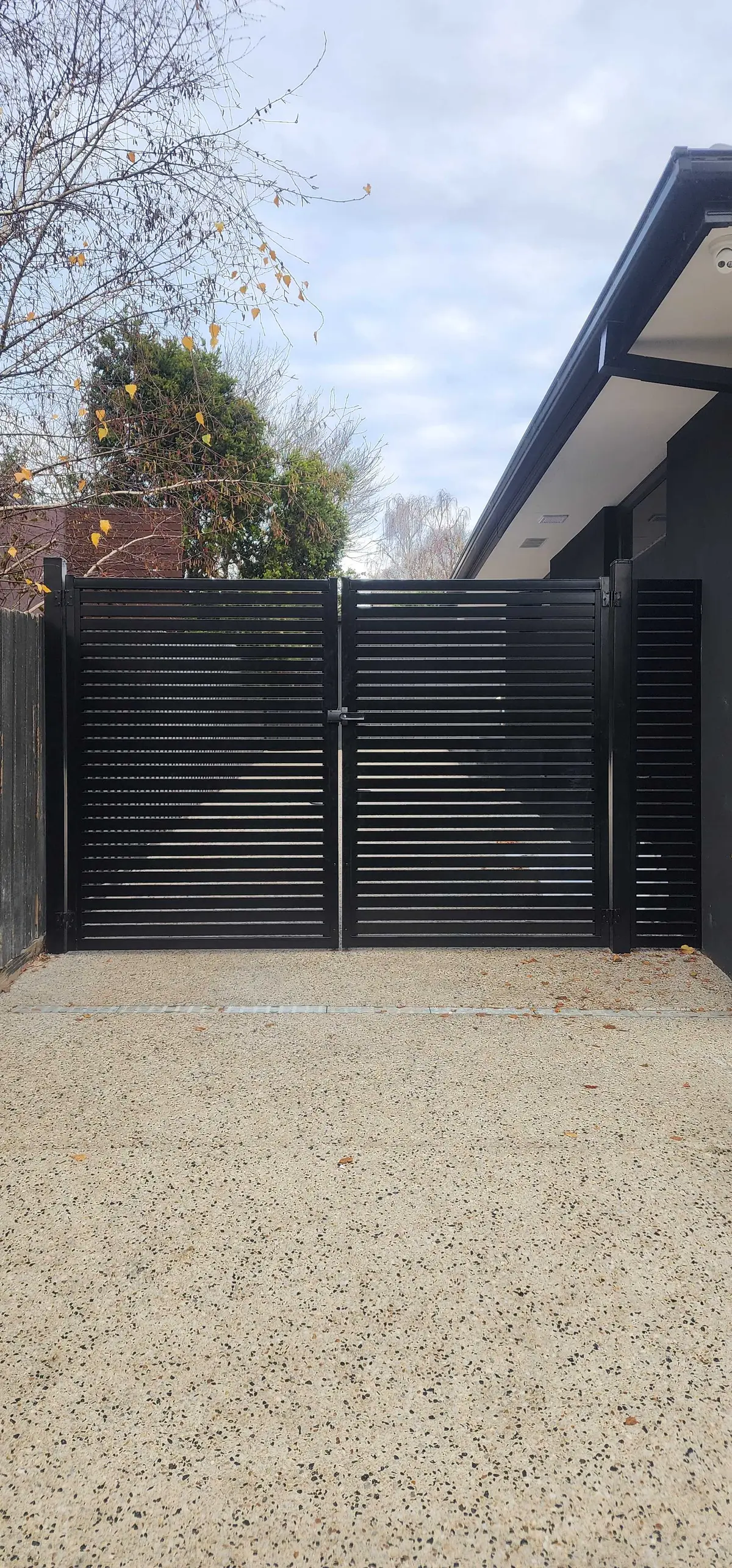 Black aluminium slat double swing driveway gates in front of a modern dark-clad home
