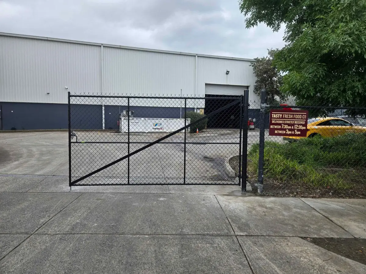 Black chain mesh swing gate with a diagonal brace across a factory driveway, seen from the street