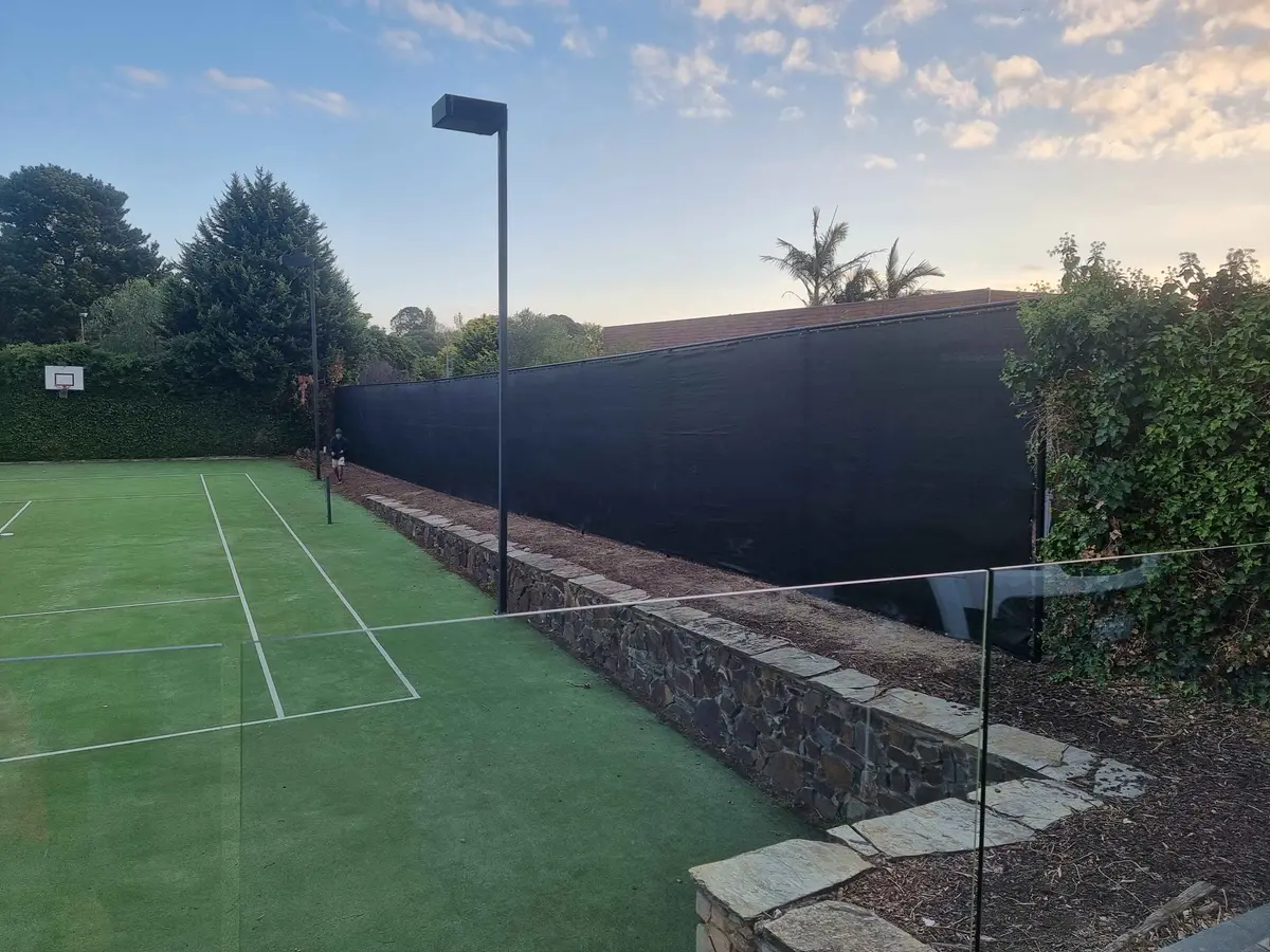 Tall black mesh tennis court fence on steel posts above a stone retaining wall